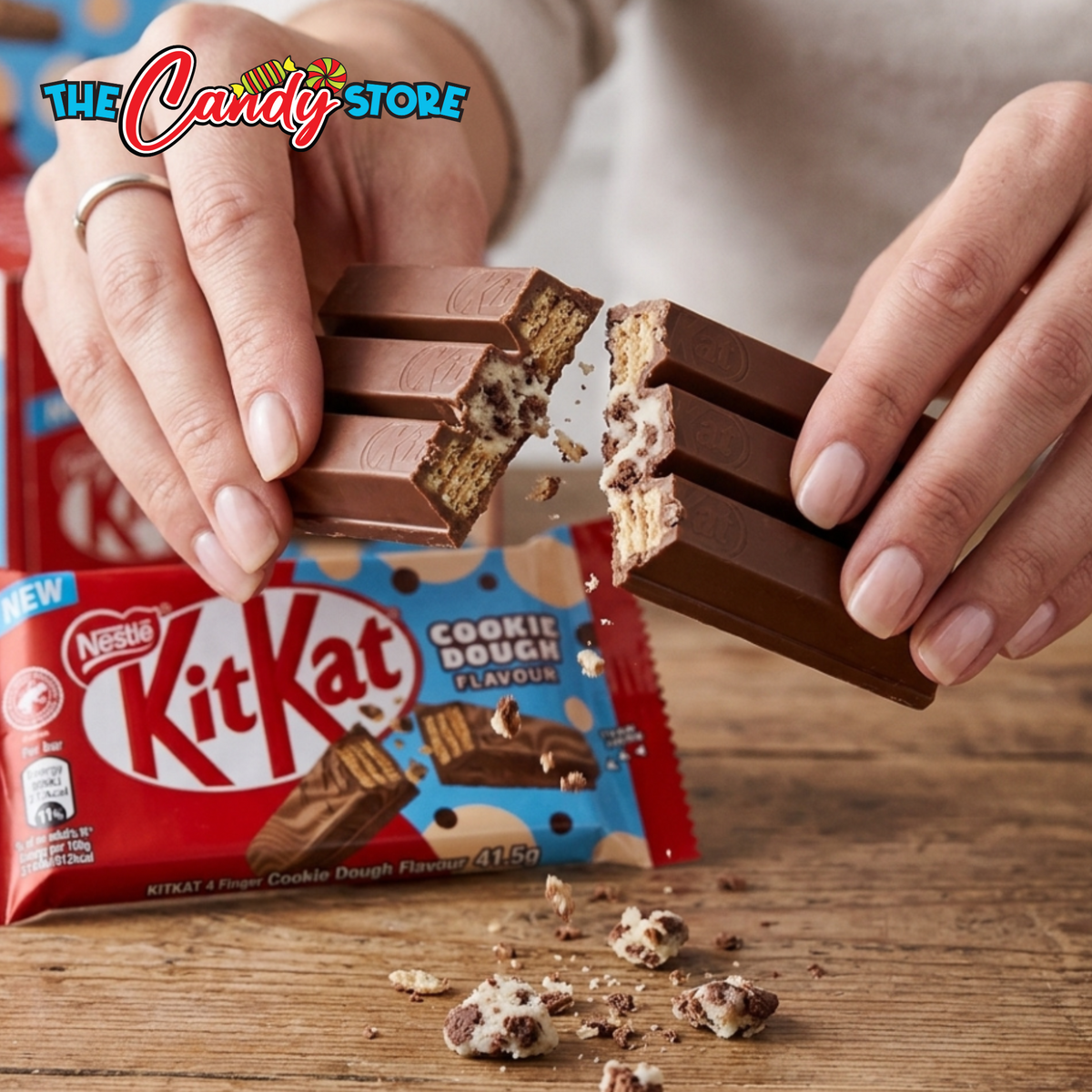 A person's hands break a KitKat Cookie Dough bar in half, showing the chocolate-coated wafers and cookie dough filling. The red and blue KitKat packaging is visible in the background on a wooden surface.