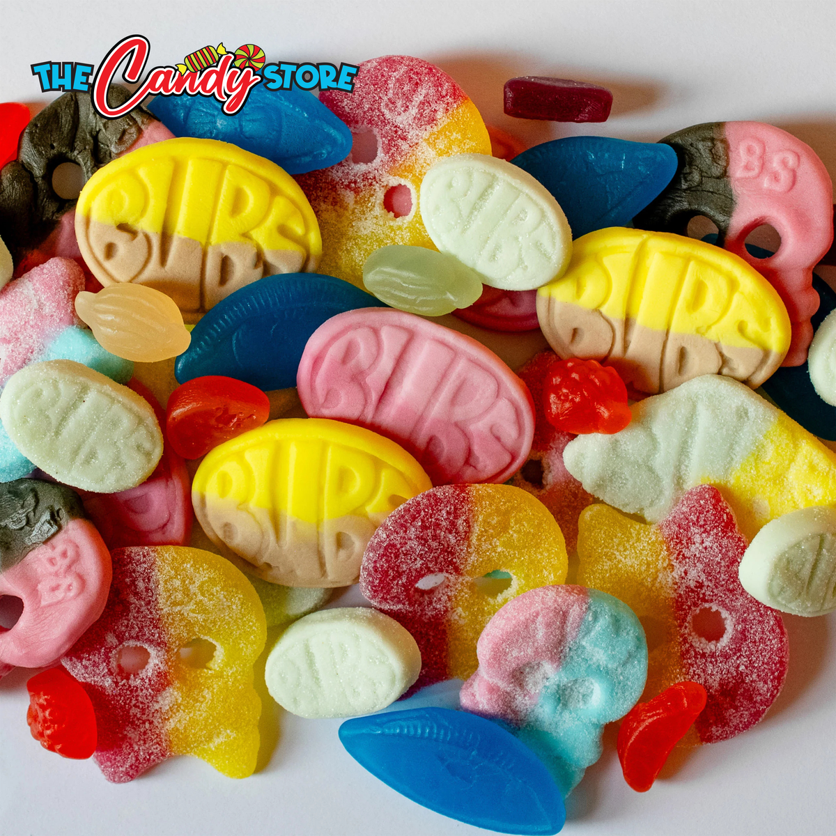 A close-up pile of assorted colorful hard candies and gummy sweets on a white background, with a 'The Candy Store' logo in the top left corner. 
